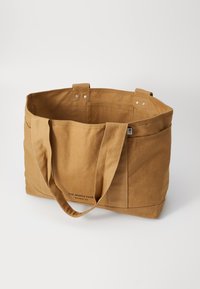 Brown canvas tote bag with two shoulder straps, spacious interior, exterior pockets, and printed branding along the bottom.