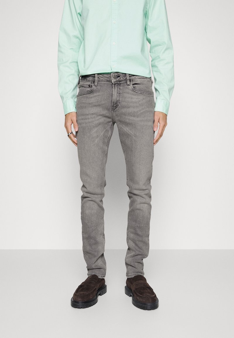 Jeans de mezclilla gris con corte slim, con cinco bolsillos y un ligero desgaste. Combinados con una camisa de botones verde claro y mocasines marrones.