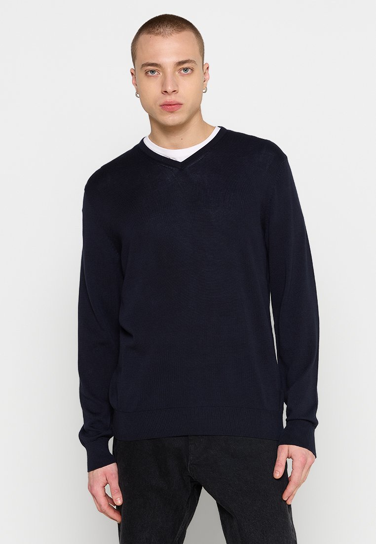 Navy-blaues V-Ausschnitt-Strickpullover mit gerippten Bündchen und Saum, aus weichem Stoff mit feiner Textur. Über einem weißen Hemd getragen.