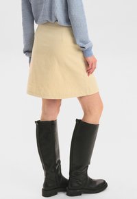 Jupe en corduroy beige longueur genou avec une coupe évasée, associée à des bottes en cuir noires hautes avec une fermeture éclair. Haut texturé bleu visible.