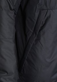 Svart puffjacka med en slät, matt yta. Har fickor med dragkedjor på sidorna och sydda sömmar, vilket visar på en funktionell design.