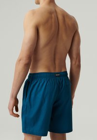 Niebieskie szorty kąpielowe z teksturowanej tkaniny, z elastycznym pasem, posiadające małą kieszonkę boczną i metkę marki z tyłu. Widok z tyłu modela.
