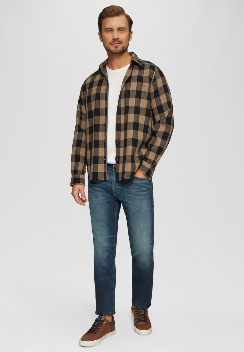 Chemise flanelle à carreaux beige et noire, t-shirt blanc en dessous, jean bleu, chaussures de sport marron avec lacets noirs, debout contre un fond uni.