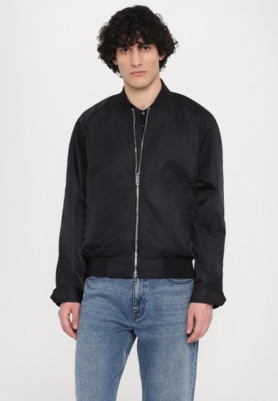 Zwarte bomberjack van gladde stof, met een rits aan de voorkant, geribbelde manchetten en een taille, gedragen over lichtblauwe jeans.