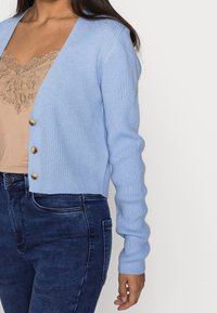 Ljusblå ribbad cardigan med långa ärmar, V-ringning och fyra dekorativa knappar. Bärs över en beige topp med spetsdetaljer och mörka jeans.