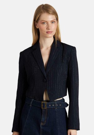 Blazer azul marino cortado con rayas verticales, cierre de un solo botón, solapas con muesca y un ligero brillo. Presenta hombreras acolchadas y un corte entallado.