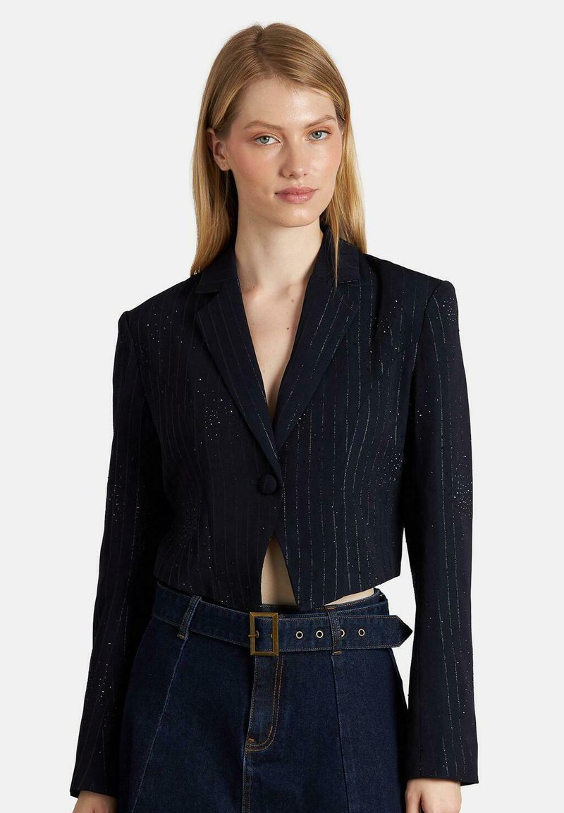 Giacca blu navy sfiancata con righe verticali, chiusura con un unico bottone, revers a punta e leggera lucentezza. Presenta spalline imbottite e vestibilità sagomata.
