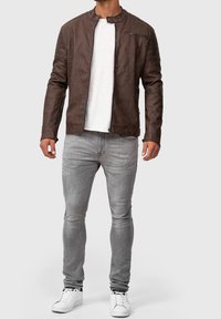 Veste en cuir marron avec un col montant, fermeture éclair à l'avant et des détails de coutures horizontales sur les épaules ; assortie avec un jean gris et des baskets blanches.
