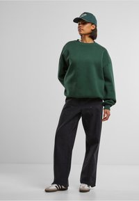 Felpa verde oversize con maniche a polsino, abbinata a pantaloni larghi blu navy. Sneakers bianche con tre strisce nere e cappellino verde.