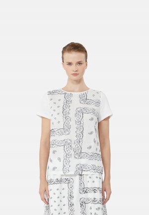 Donna che indossa un top bianco a maniche corte e una gonna con stampa paisley bandana nera, in piedi contro uno sfondo chiaro e uniforme.