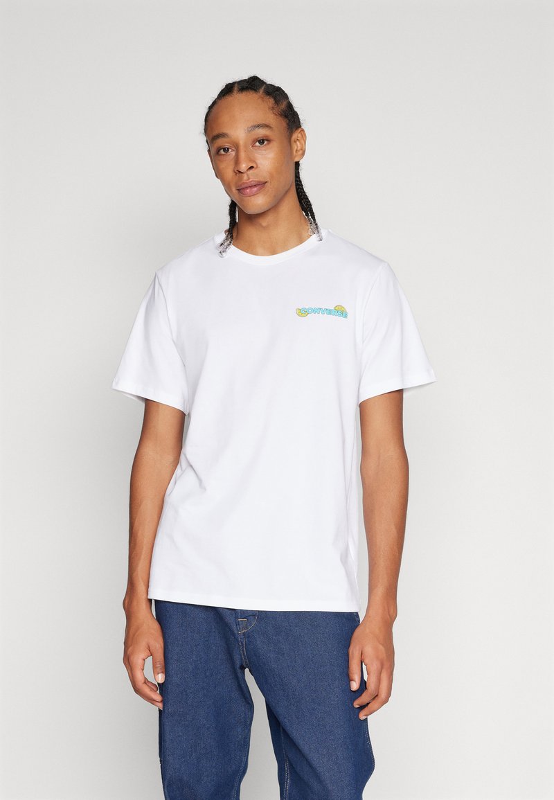 Camiseta de algodón blanca con mangas cortas, que presenta un pequeño logo multicolor de "Converse" en el lado izquierdo del pecho. Diseño clásico de cuello redondo.