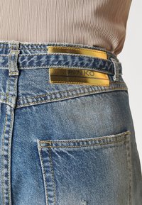 Vue arrière d’un jean en denim bleu avec des coutures jaunes et une étiquette dorée portant la mention "PINKO" sur la ceinture, porté avec un top beige côtelé.