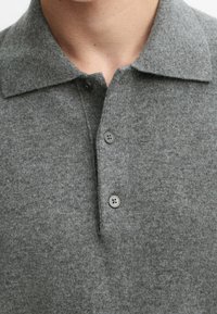 Nahaufnahme einer Person, die ein graues gestricktes Poloshirt mit drei Knöpfen trägt, von denen zwei geschlossen und einer am Kragen offen ist.