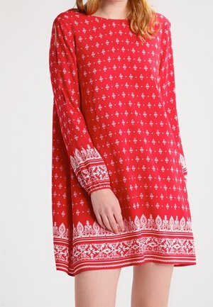 Femme portant une robe rouge à manches longues avec des motifs géométriques et floraux blancs sur les manches et l'ourlet, debout sur un fond uni.