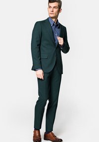Ciemnozielony garnitur dopasowany z klapami, zestawiony z niebieską koszulą na guziki. Brązowe skórzane buty dopełniają całość.