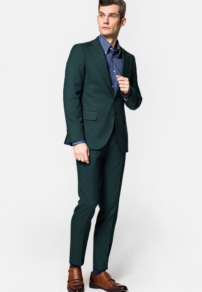 Ciemnozielony garnitur dopasowany z klapami, zestawiony z niebieską koszulą na guziki. Brązowe skórzane buty dopełniają całość.