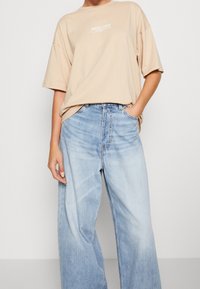 Béžové oversize tričko s bílým nápisem "PACIFIC COAST", spárované se světle modrými volnými džínami s opotřebovaným vzhledem a vysokým pasem.