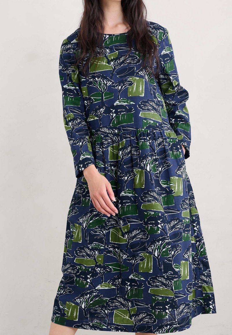 Mörkblå långärmad klänning med grön och vit botanisk tryck, med skiktad kjol och två sidofickor. Bomullstextur.