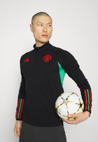 Jersey de manga larga negra con el logo rojo del Manchester United, paneles laterales verdes y acentos de rayas multicolores en las mangas. Sosteniendo un balón de fútbol.