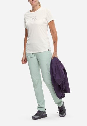 Femme en pantalon vert clair et t-shirt blanc tenant une veste violette pliée, portant des chaussures de randonnée grises, debout sur un fond blanc.