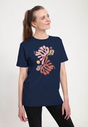 Kvinde med brun hestehale iført en mørkeblå T-shirt med koralinspirerede abstrakte former og tekst på en ensfarvet lys baggrund.
