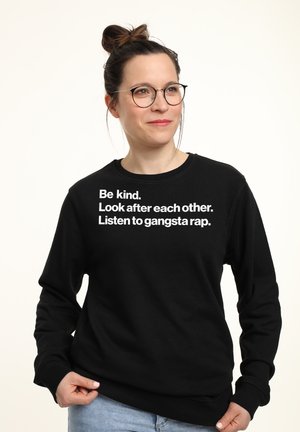 Sweatshirt noir avec un col rond. Il présente un texte blanc : "Soyez gentils. Prenez soin les uns des autres. Écoutez du gangsta rap." Matière en coton doux.