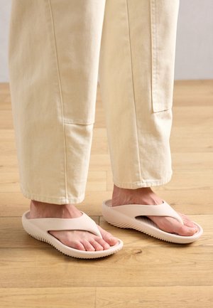 Rózsaszín gumi papucs, keresztpántos kialakítással, bordázott talppal és enyhe ív támogatással, fehér bőszárú nadrággal viselve egy fa padlón.
