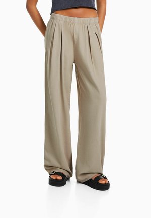 Mujer con pantalones anchos de tiro alto beige con pliegues combinados con sandalias negras con hebilla y un top corto oscuro.