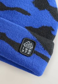 Modrá pletená čiapka s čiernym maskáčovým vzorom, s prehnutým okrajom a obdĺžnikovou čiernou nášivkou s horským logom a rokom "1982."