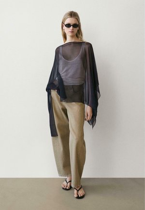 Donna con capelli biondi lunghi che indossa occhiali da sole neri, una maglia nera trasparente sopra una canottiera grigia, pantaloni beige a gamba larga e infradito nere.