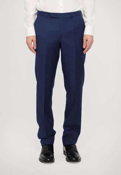 Pantalons taillés marine en tissu fin, avec une taille plate, des jambes droites et un détail de pli, associés à des chaussures de ville noires.
