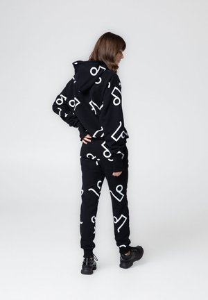 Kobieta stojąca bokiem, ubrana w czarną bluzę z kapturem i spodnie z białymi abstrakcyjnymi geometrycznymi wzorami, czarne sportowe buty, na jasnym, jednolitym tle.