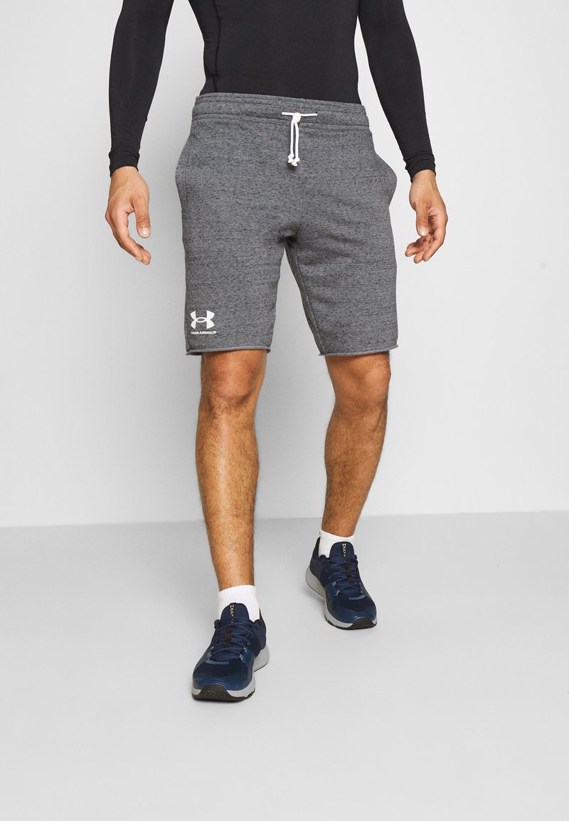 Homme portant un short gris Under Armour, un haut à manches longues noir, des chaussettes blanches et des chaussures de sport bleues, debout sur un sol gris.