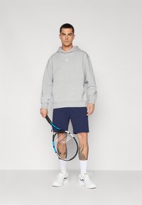 Grå hættetrøje med et logo på forsiden, parret med marineblå shorts. Figuren holder en sort tennisracket med blå detaljer, iført hvide sko og sokker.