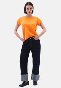 Oranžové bavlněné tričko s krátkým rukávem, spárované s tmavými džínami s kontrastními detaily na manžetách a zlatým prošíváním. Černé boty.