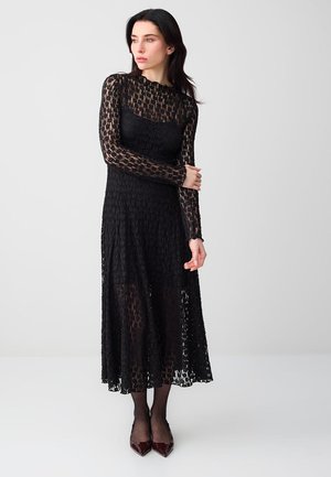 Frau mit dunklem Haar, die ein langärmeliges, schwarzes Spitzen-Midikleid und burgunderrote spitze Absätze trägt, steht vor einem schlichten grauen Hintergrund.