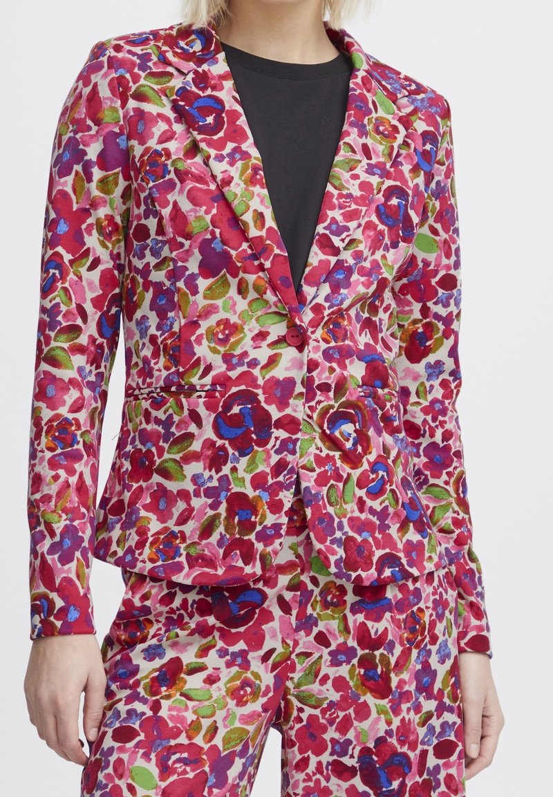 Femme portant un blazer à motif floral rose, rouge, bleu et vert avec un pantalon assorti sur une chemise noire.