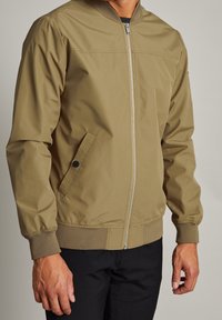 Chaqueta bomber color caqui, hecha de un material suave y ligero, con cremallera frontal, bolsillos laterales y puños y dobladillo acanalados.