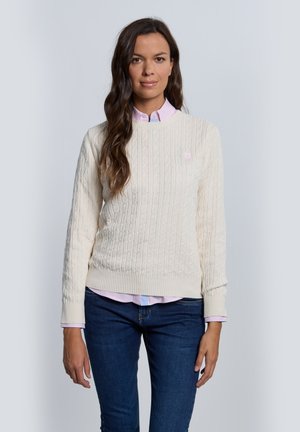 Suéter de punto grueso color crema con puños y dobladillo acanalados, que presenta un pequeño emblema. Debajo, se observa una camisa de cuello de color rosa claro.
