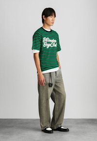 Grøn- og sortstribet T-shirt med hvide accenter og logo, båret over en hvid langærmet skjorte. Sættet kombineret med løse grå jeans og sorte sko.