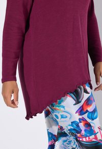 Bordeauxfarbene Langarmbluse mit gewelltem Saum, kombiniert mit bunten Leggings im Blumenmuster in den Farben Blau, Pink und Weiß.