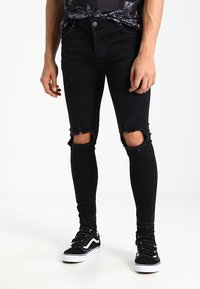 Jean skinny noir déchiré avec des ouvertures déchirées aux genoux, associé à des baskets noir et blanc. Le matériau semble être du denim avec une texture lisse.