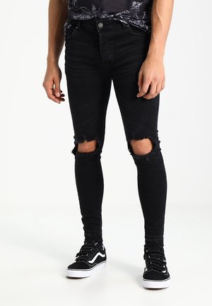 Jean skinny noir déchiré avec des ouvertures déchirées aux genoux, associé à des baskets noir et blanc. Le matériau semble être du denim avec une texture lisse.