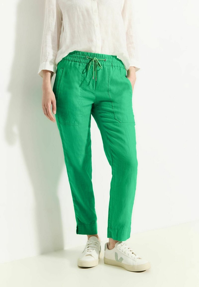 Personne portant un pantalon vert vif à cordon, une chemise blanche boutonnée et des baskets blanches, debout contre un mur blanc.