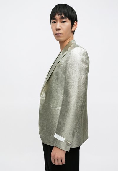 Marynarka w kolorze metalicznego złota z teksturą połyskującą, z wyciętymi klapami i dopasowanym krojem. Na mankiecie znajduje się biała metka z napisem "Versace".