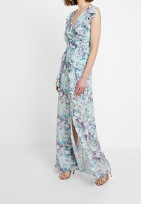 Femme portant une robe sans manches, longue jusqu'aux chevilles, de couleur bleu clair avec des fleurs, des volants et une fente à l'avant, associée à des sandales à talons nus.