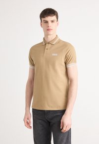 Braunes Poloshirt mit klassischem Kragen, ausgestattet mit weißen gestreiften Akzenten an den Ärmeln und einem Markenpatch auf der Brust. Tragen mit dunklen Jeans.