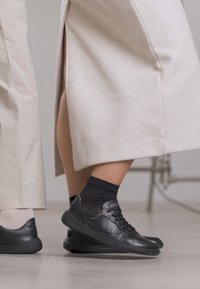 Dos personas con zapatillas negras paradas cerca; una con pantalones beige claro y la otra con falda beige y calcetas negras.