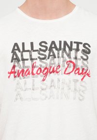 Λευκό πουκάμισο με στρωματοποιημένο κείμενο «ALLSAINTS» σε μαύρο και γκρι και «Analogue Days» με κόκκινη γραφή πάνω στο στήθος.