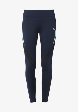 Kobieta idąca, ubrana w biały T-shirt, granatowe legginsy z jasnoniebieskimi i czarnymi ukośnymi panelami oraz białe buty sportowe na jednolitym tle.